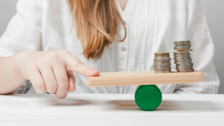 Hand balanciert Münzstapel auf einer Wippe – sinnbildlich für die Gewinnermittlung als Gleichgewicht von Einnahmen und Ausgaben.